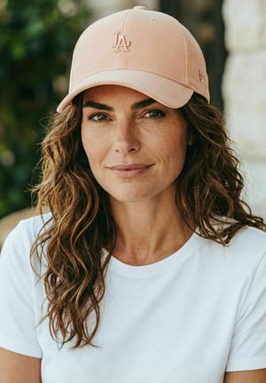 Femme aux cheveux bruns ondulés, portant une casquette beige LA et un t-shirt blanc, souriant doucement devant un arrière-plan naturel flou.