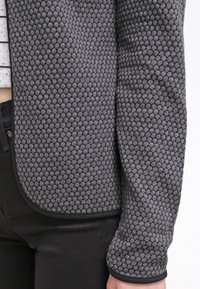 Grauer strukturierter Pullover mit Wabenmuster, runder Ausschnitt und schwarzer Kante entlang der Säume und Ärmel.