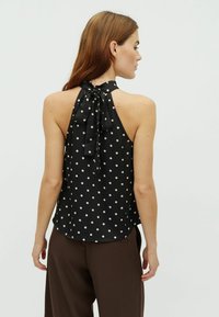 Čierny halter top s bodkami a viazaním na chrbte, s voľným strihom a hladkým povrchom. Nosený s hnedými nohavicami.