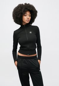 Čierna bunda na zips s rebrovanou textúrou, vybavená vysokým golierom a vyšitým logom. V kombinácii s čiernymi nohavicami s bočnými vreckami.