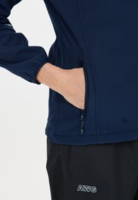 Marinblå softshell-jacka med en dragkedjeficka, elastiska ärmslut och logodetaljer. Har en slät yta och en figurnära silhuett.