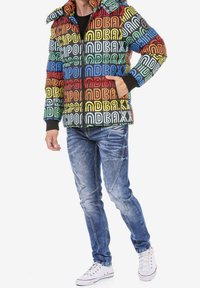 Kleurrijk pufferjack met geometrisch tekstpatroon in rood, blauw, groen en geel. Gecombineerd met versleten blauwe jeans en witte sneakers.