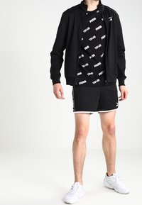 Veste de sport noire avec un col haut, accompagnée d'un T-shirt graphique noir et d'un short noir. Des baskets blanches complètent le look.