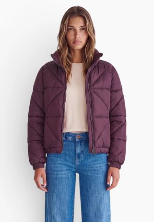 Femme portant une veste matelassée violette sur une chemise blanche et un jean bleu, debout devant un fond blanc uni.