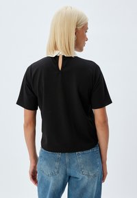 Zwarte t-shirt met korte mouwen en een ronde halslijn, voorzien van een klein keyhole-detail aan de achterkant, gecombineerd met lichtblauwe, high-waisted jeans.