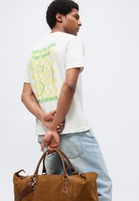 Άνδρας με ανοιχτόχρωμο τζιν και λευκό μπλουζάκι με σχέδιο λεμονιού κρατώντας καφέ σουέντ τσάντα ταξιδιού πίσω από την πλάτη του, βλέποντας πλάγια σε λευκό φόντο.