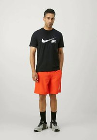 Camiseta de algodón negra con el logo blanco de Nike, combinada con pantalones cortos naranja brillante, calcetines negros y zapatillas deportivas grises con un diseño texturizado.