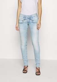 Helle blaue enge Jeans mit ausgewaschenen Stellen, detaillierten Fronttaschen, Knopfverschluss und einem eleganten Design, kombiniert mit schwarzen Riemchensandalen.