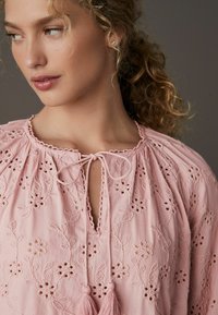 Blusa rosa ricamata con motivi floreali, scollatura a chiave, chiusura a cravatta e dettagli con frange. Tessuto morbido con finiture strutturate.