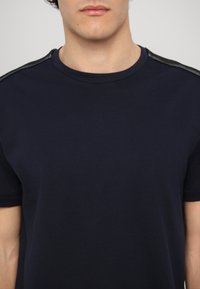 T-shirt de algodão azul-marinho com mangas curtas, decote redondo e detalhes texturados em preto nos ombros. Tecido suave, corte casual.