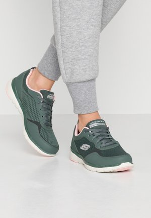 Osoba obuvající zeleno-růžové tenisky Skechers s šedými tepláky, stojící na hladkém bílém povrchu.