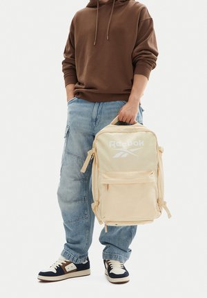 Persona con sudadera con capucha marrón y vaqueros azules sosteniendo una mochila beige de Reebok por el asa superior, de pie sobre fondo blanco.