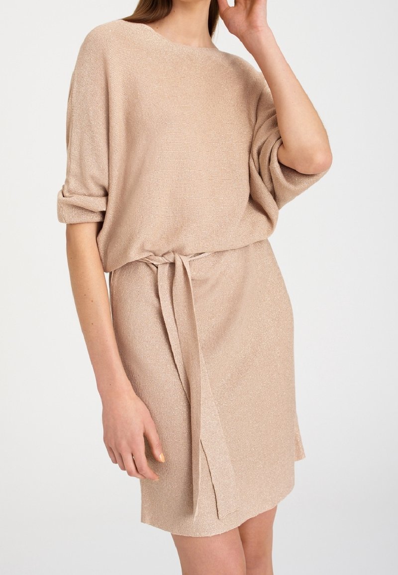 Robe en maille beige avec une coupe ample, des manches courtes et une ceinture à nouer à la taille. Présente un léger scintillement et une fente latérale pour plus de détails.