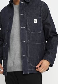 Džínová bunda v tmavě indigové barvě s bílým šitím, vybavená náprsní kapsou a knoflíkovými zapínáními. Nošena přes šedou mikinu.