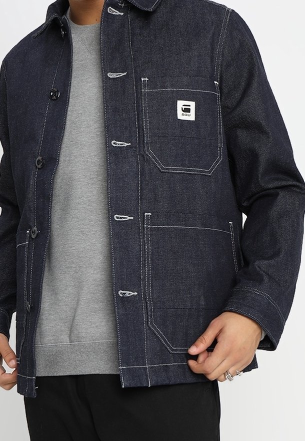 Džínová bunda v tmavě indigové barvě s bílým šitím, vybavená náprsní kapsou a knoflíkovými zapínáními. Nošena přes šedou mikinu.