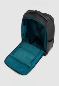Mochila negra con interior en color azul verdoso, compartimento abierto que muestra correas ajustables y bolsillos de malla con cremallera en la solapa interior.