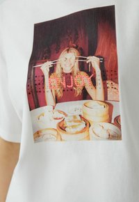 Witte T-shirt met een kleurrijke fotoafdruk van een vrouw die eetstokjes boven een tafel met dim sum houdt. Tekst "ENJOY" in roze strass.