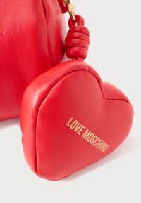 Raudona odinė rankinė su tekstūruotu paviršiumi ir širdelės formos žavesiu. Auksinė "LOVE MOSCHINO" logotipas ant žavesio. Yra mazgo detalė ant dirželio.