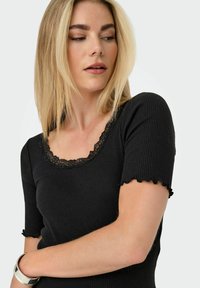 Femme blonde regardant sur le côté, portant un haut côtelé noir avec un col en dentelle et des manches courtes festonnées, bracelet argenté au poignet.