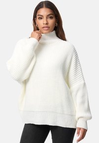 Dik gebreide witte trui met een hoge hals, oversized mouwen en een geribbelde zoom. De textuur is zacht en ruimvallend.