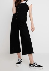 Svart denim jumpsuit med vida ben, framknäppning och fransad nederkant. Bärs över en vit T-shirt och matchas med svart och vita sneakers.