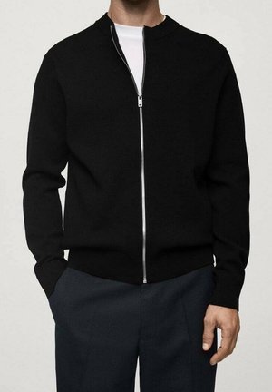 Homme portant un pull noir à fermeture éclair sur une chemise blanche et un pantalon foncé, avec une main dans la poche sur un fond uni.