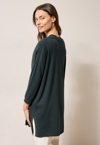 Mörkgrön, oversized stickad tunika med rundad halsringning, trekvartsärmar och sidofrånskärningar. Slät textur, avslappnad design.