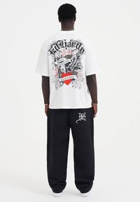 Vit grafisk t-shirt med djärva svarta och röda mönster på ryggen, kombinerad med svarta byxor som har en vit logodetalj.