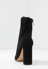 Bottine en daim noir avec un talon bloc haut, une fermeture éclair à l'arrière et un design incurvé. Texture lisse et finition naturelle sur la matière supérieure.