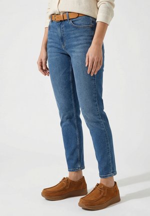 Person trägt blaue Slim-Fit-Jeans, braunen Gürtel, beigen Pullover und braune Wildlederschnürschuhe und steht vor hellem Hintergrund.