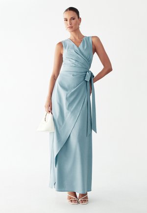 Vrouw draagt een lichtblauwe mouwloze wikkelmaxi-jurk, houdt een kleine witte handtas vast en draagt witte sandalen met bandjes tegen een witte achtergrond.