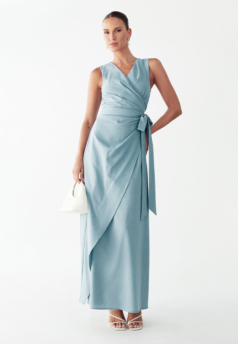 Naine kannab helesinist varrukateta maksikleiti, hoiab käes väikest valget käekotti ja kannab valgeid rihmikuid kontsakingi, valgel taustal.