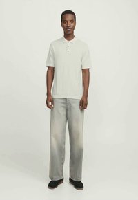 Mann som står fremovervendt iført en hvit kortermet polo-skjorte, lysegrå vide jeans og svarte sko.