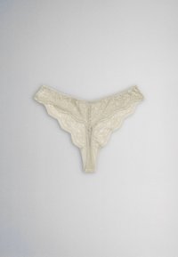 String en dentelle beige avec motifs floraux et bords festonnés, posé à plat sur un fond gris clair.