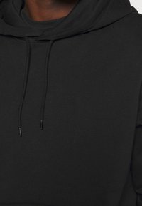 Zwarte hoodie van een zachte, gestructureerde stof. Kenmerken zijn een grote kap, koorden en een relaxte pasvorm. Geen zichtbare logo's of patronen.