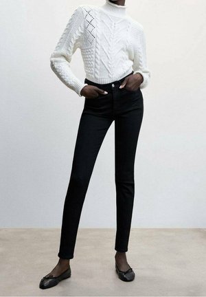Maglione bianco a collo alto in lavorazione a treccia con dettagli a pattern, abbinato a jeans neri a vita alta e ballerine nere.