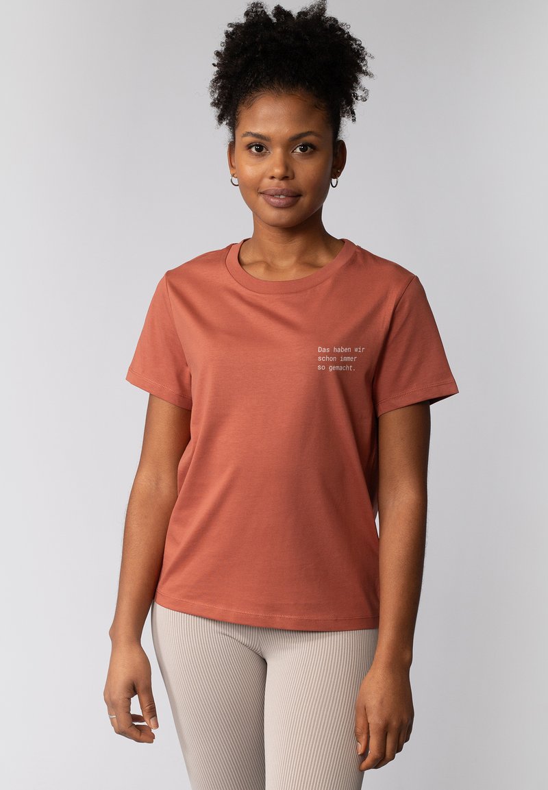 T-shirt en coton couleur rouille avec des manches courtes, col rond et détail de texte dans une couleur contrastante à l'avant. Texture lisse.