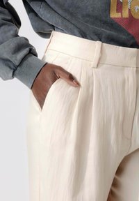 Pantalons plissés beiges en tissu léger, dotés de poches latérales et associés à un sweat-shirt gris avec des manches texturées.