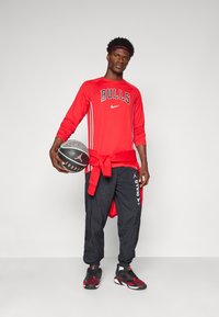 Rød langærmet skjorte med "BULLS" logo og Nike swoosh, parret med sorte træningsbukser med logo-print, og sorte sneakers mens der holdes en basketball.