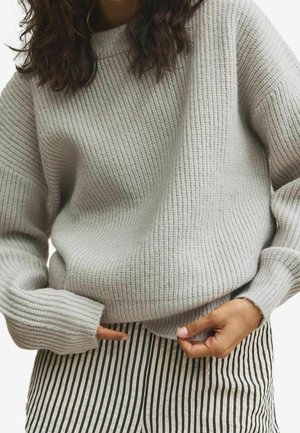 Femme portant un pull en tricot côtelé gris clair et un pantalon à rayures verticales noires et blanches, les mains ajustant l'ourlet du pull.