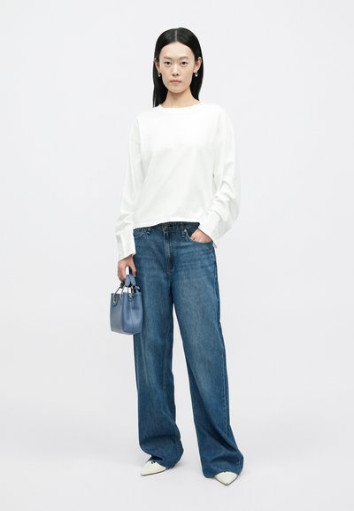 Biała koszula z długim rękawem, luźne niebieskie jeansy i jasnoniebieska torebka; białe szpiczaste buty dopełniają casualowy strój. Jednolite tło.