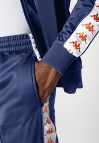 Marine joggedress med glidelåsjakke og bukser. Hvite sidepaneler med oransje logoakenter. Glatt, lett materiale.