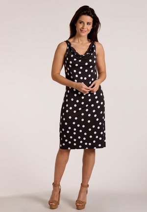 Vrouw staat, draagt een mouwloze zwarte jurk tot op de knie met witte polka dots en bruine sandalen met hoge hakken, handen losjes samen.