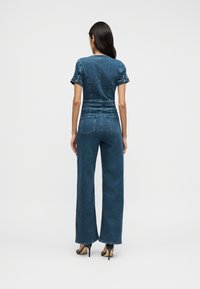 Ολόσωμη φόρμα από τζιν σε σκούρο μπλε, με κοντά μανίκια και εφαρμοστή μέση. Σχεδιασμός με φαρδιά πόδια, πίσω τσέπες και διακριτικές ραφές.