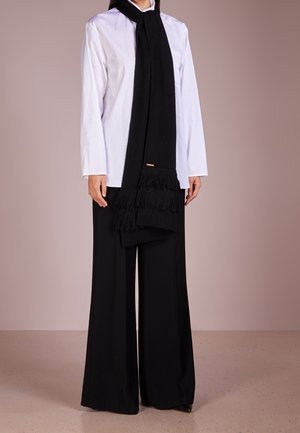 Femme portant une chemise blanche à manches longues, un pantalon noir à jambes larges, et une longue écharpe noire à franges drapée autour du cou.