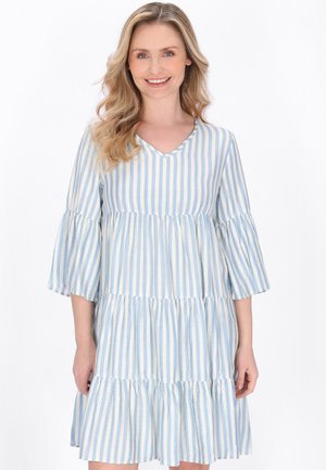 Femeie zâmbitoare cu păr blond ondulat, purtând o rochie lejeră cu volane, cu dungi verticale alb-albastre și mâneci evazate de trei sferturi.