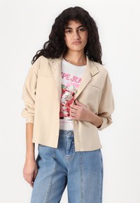 Camisa de botones beige hecha de tejido ligero, con cuello y dos bolsillos en el pecho, superpuesta a una camiseta gráfica, combinada con jeans azules.