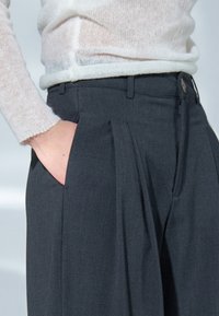 Harmaat, laskostetut housut, joissa on sivutaskut, vyölenkit ja hienovarainen tekstuuri, yhdistettynä kevyeseen, läpikuultavaan valkoiseen neulepuseroon.