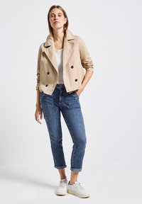 Beige Doppelreiher-Jacke mit schwarzen Knöpfen, kombiniert mit dunkelblauen Jeans mit aufgerolltem Saum und weißen Sneakers, vor einem hellen Hintergrund.