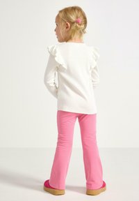Ung pige med blondt hår i en knold iført en hvid langærmet top med skulderpuf, lyserøde bukser og lyserøde fluffy slippers, vendt mod venstre.